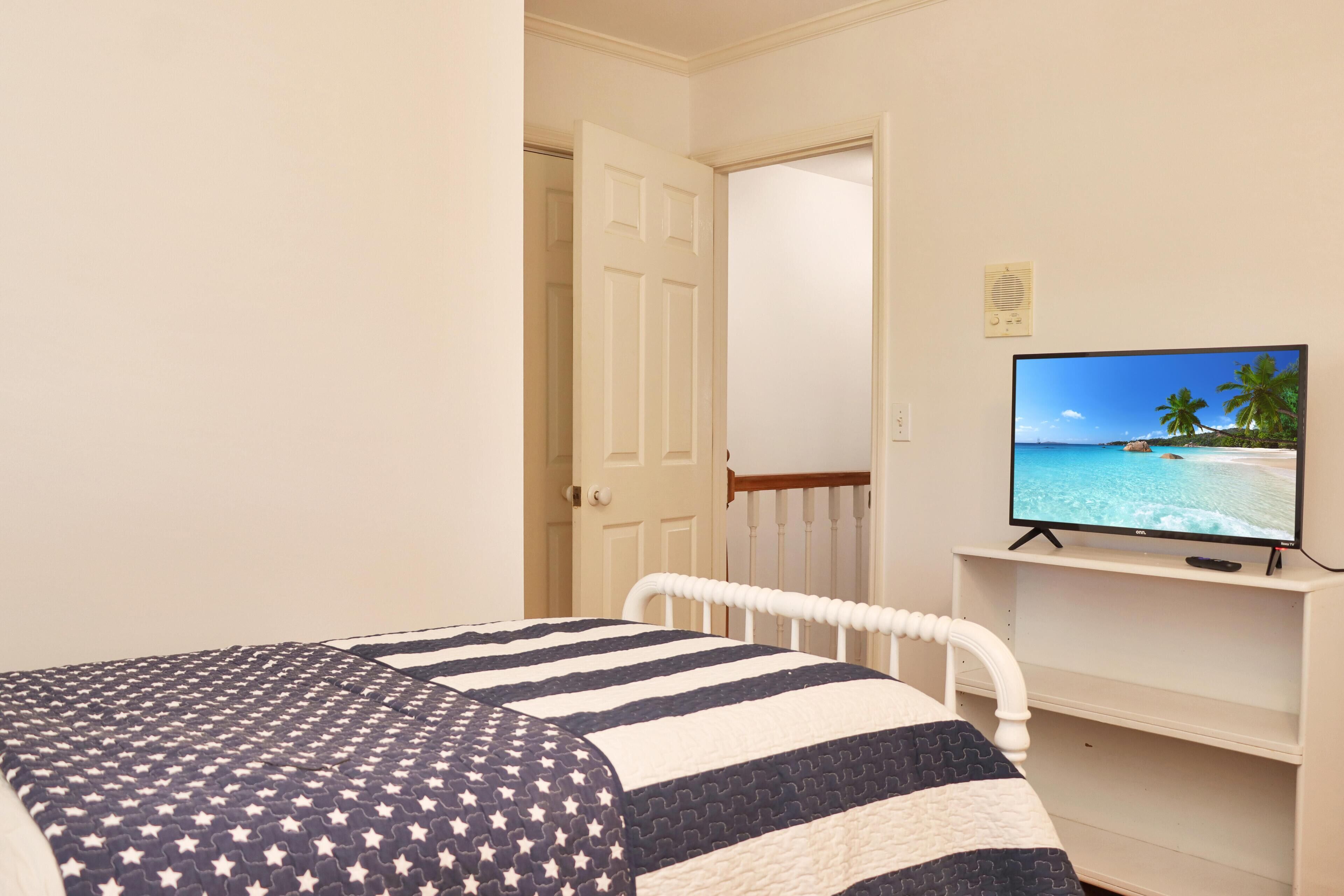 Une chambre à coucher avec un lit, un téléviseur diffusant une scène de plage, et une porte donnant sur une autre pièce.
