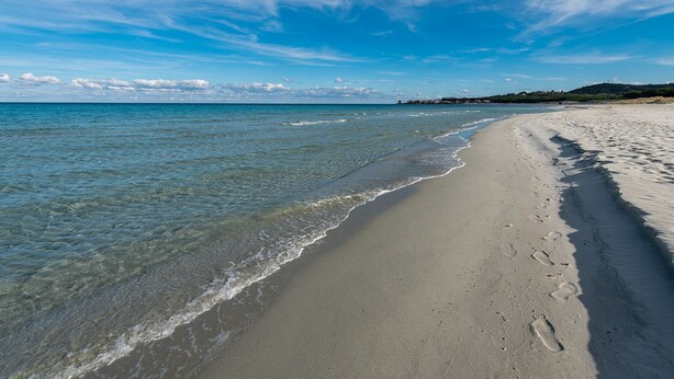 Een strand met helder water, een zandstrand en een verre kustlijn met gebouwen.