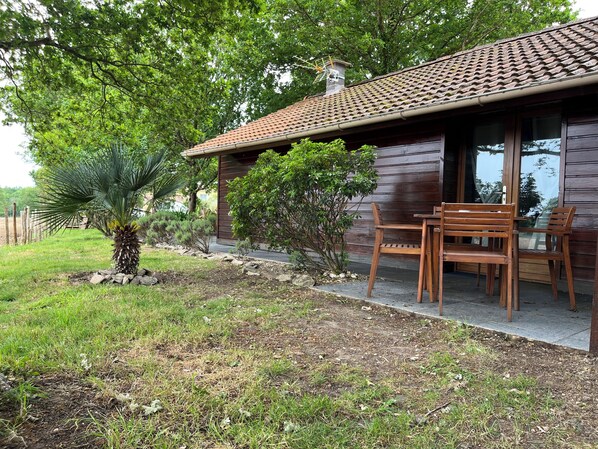 Une maison en bois avec un toit en tuiles, une terrasse avec une table et des chaises en bois, et un petit palmier.