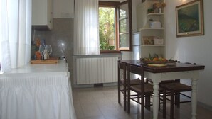 Un comedor con mesa, sillas, un radiador, una ventana con cortinas y una estantería con utensilios de cocina.