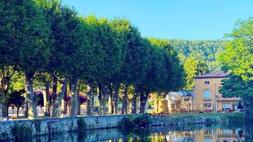 A tranquil riverside scene with a row of trees reflecting in the water, a stone wall, and a building with a tiled roof in the background.