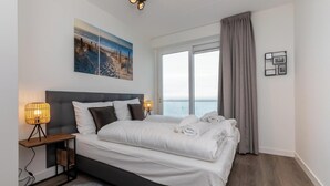 A modern hotel room with a large bed, a nightstand with a lamp, a view of the sea through large windows, and a painting on the wall.
