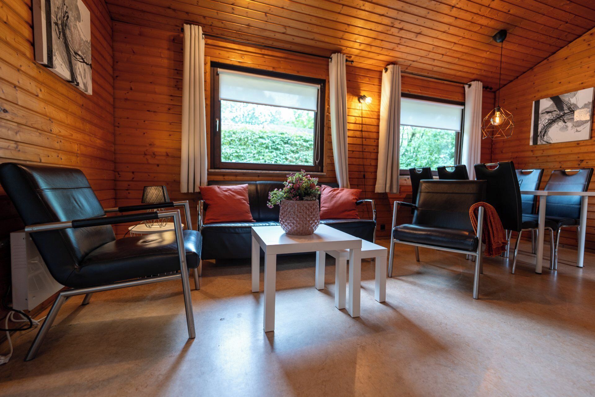 A cozy cabin interior with wooden walls, a sofa, two armchairs, a small table with a plant, and a dining area with chairs.