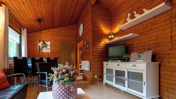 A cozy cabin interior with wooden walls, a dining area with chairs and a table, a TV, and a cabinet with drawers. - Type 5p - Two Bedroom Chalet, Sleeps 5 (Uelsen)