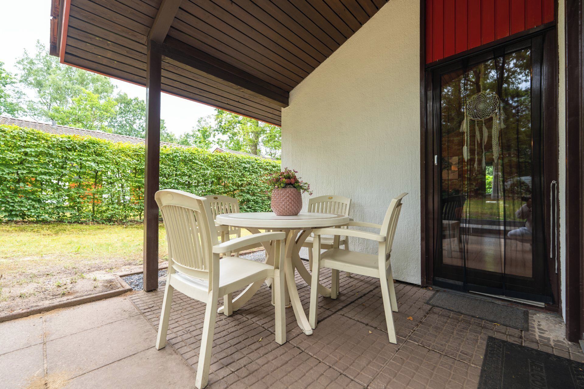 A covered patio area with a table and four chairs, a potted plant, and a glass door leading to a garden.