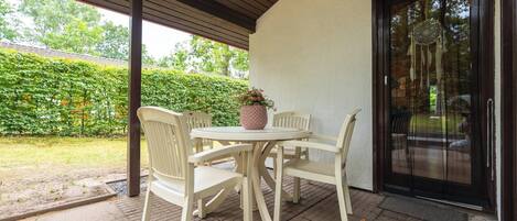 A covered patio area with a table and four chairs, a potted plant, and a glass door leading to a garden.