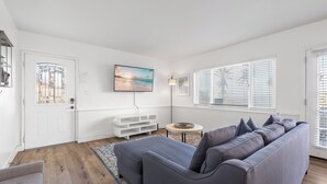 A modern living room with a grey sofa, a flat-screen TV, a white door with a decorative glass panel, and a large window with blinds. - Huge Outdoor Space/Steps to Beach & Wedding Bowl (La Jolla)
