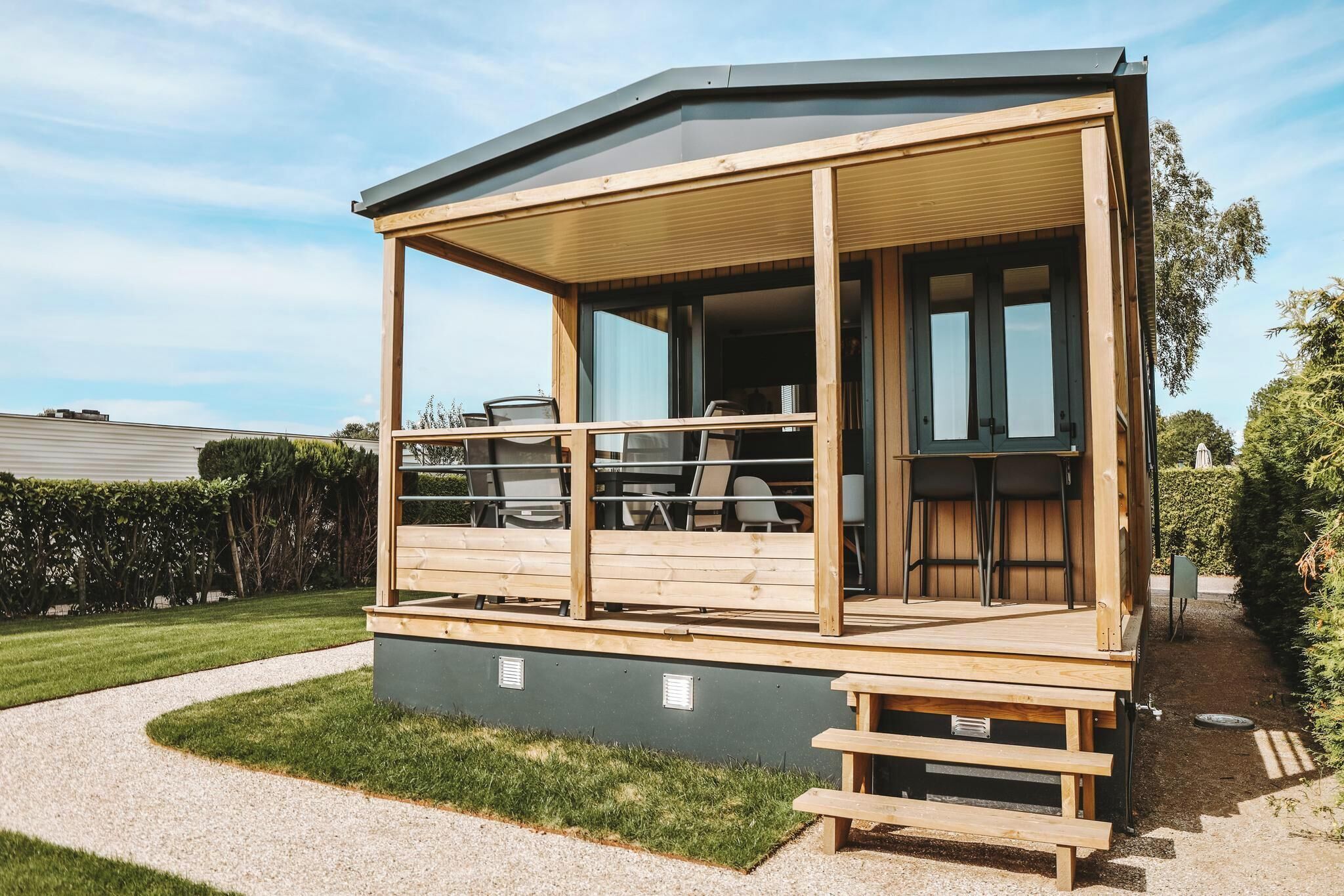 Una terraza de madera con mobiliario exterior y una zona cubierta, rodeada de un jardín cuidado.