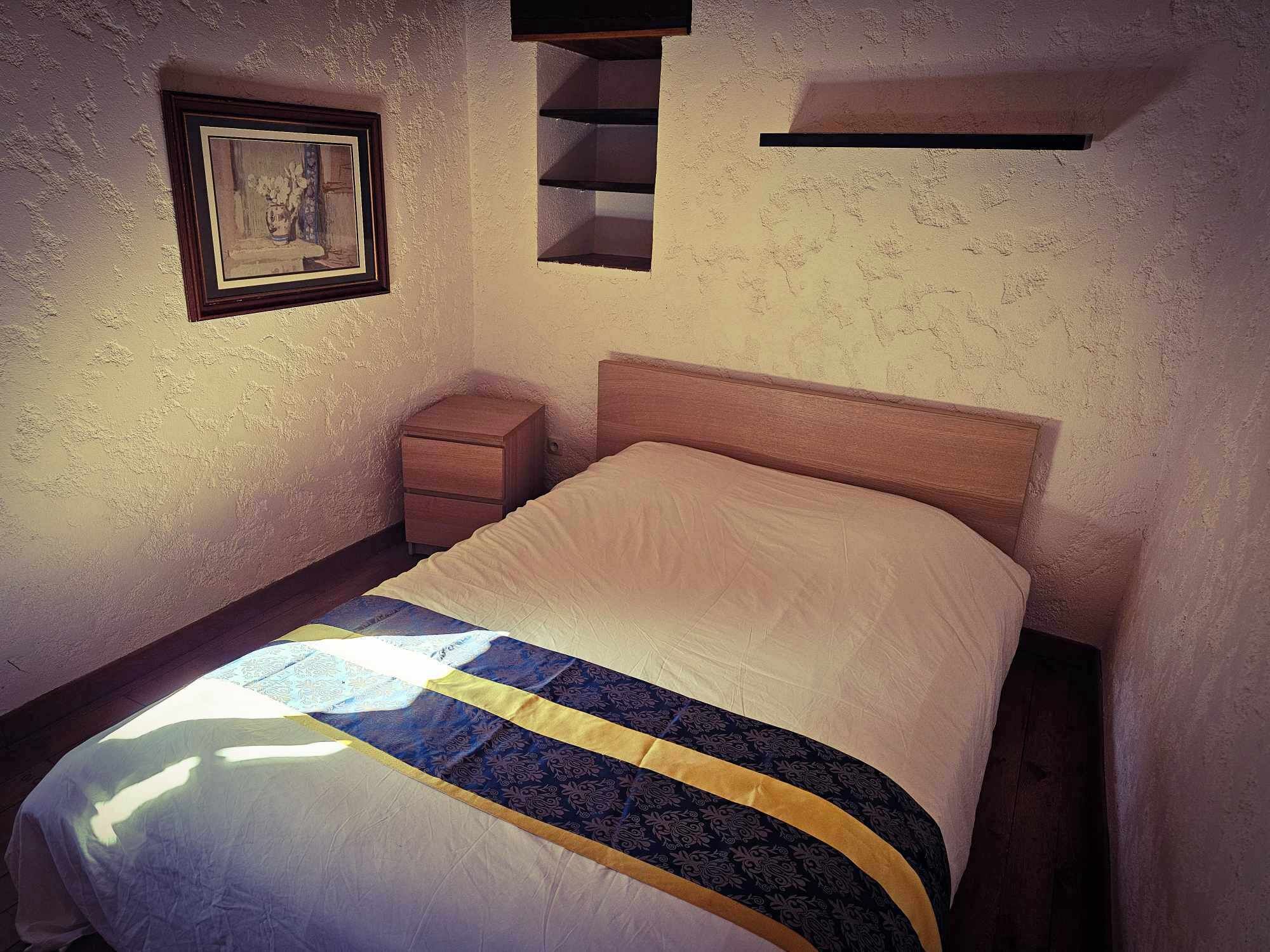 A bedroom with a bed, a nightstand, a framed picture on the wall, and built-in shelves.