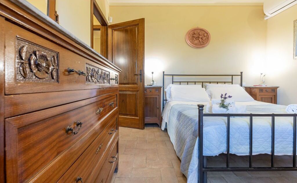 Quarto com cômoda de madeira, cama com roupa de cama branca, mesa de cabeceira e relógio de parede.