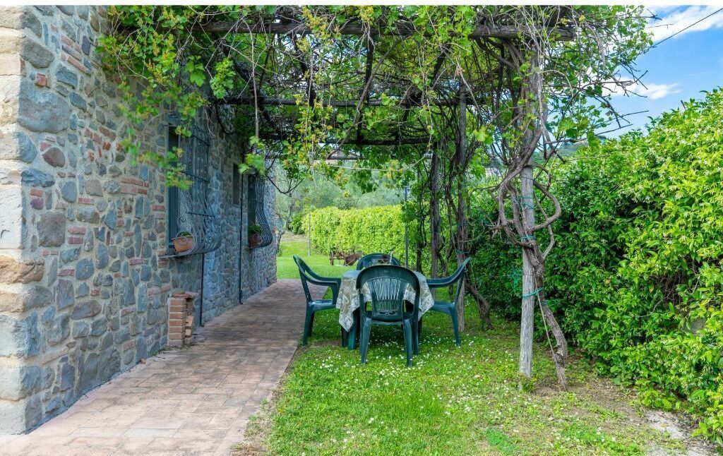 A stone wall with a wooden pergola, a grassy area with a table and chairs, and a hedge in the background.