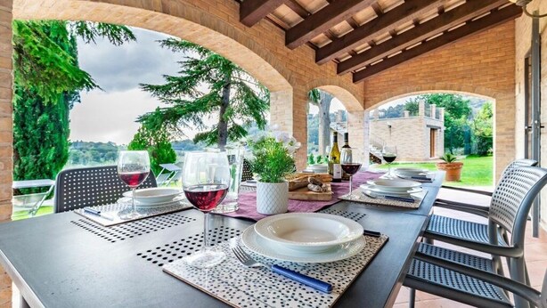 An outdoor dining area with a brick archway, a table set for two with wine glasses and plates, and a view of the surrounding greenery and hills.