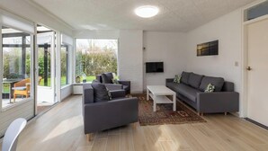 Ein modernes Wohnzimmer mit einer grauen Sofagarnitur, einem weißen Couchtisch und einem gemusterten Teppich. An der Wand ist ein Fernseher angebracht und ein großes Fenster bietet einen Blick ins Freie.