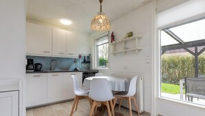Eine moderne Küche mit weißen Schränken, einem Esstisch mit weißen Stühlen, einer Pendelleuchte und einem Fenster mit Blick auf einen Garten.