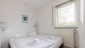 Ein sauberes, weißes Schlafzimmer mit einem Bett, zwei Kissen, einem zusammengerollten Handtuch, einer Heizung, einem Fenster mit Jalousien und einem gerahmten Bild an der Wand.