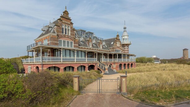 Ein großes, verziertes Gebäude mit mehreren Balkonen und einem Turm, umgeben von grünen Feldern.
