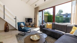 A modern living room with a grey sofa, a blue armchair, a TV, and a large window offering a view of a garden and buildings. - Holidayhouse - Lepelblad 42 | Nieuwvliet-Bad (Nieuwvliet)