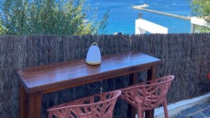 A wooden table with two pink chairs and a white lamp on a balcony overlooking a coastal view with the sea and distant buildings. - Castell Mar 1012 - Studio Apartment, Sleeps 2 (Roses)