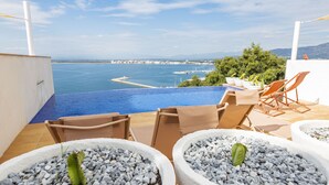 Une piscine à débordement offrant une vue sur la mer et les bâtiments lointains, entourée de chaises longues et de plantes en pot.