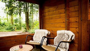 Una cabaña de madera con dos sillones cubiertos de pieles, una pequeña mesa redonda con un vaso y un objeto rosa, y vista a un bosque a través de un amplio ventanal.