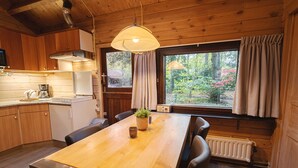 Interior de una cabaña de madera con cocina, mesa de comedor, sillas, una planta en maceta y una ventana con cortinas.