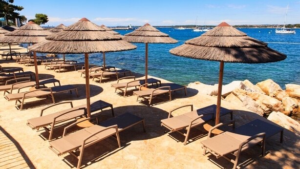A beachside lounge area with sun loungers and thatched umbrellas, overlooking a calm sea with boats.