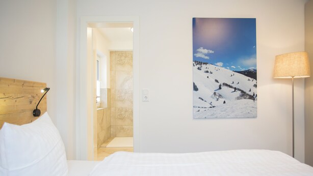 Ein Hotelzimmer mit einem Bett, einem Gemälde einer verschneiten Landschaft, einer Lampe und einem hölzernen Kopfteil.