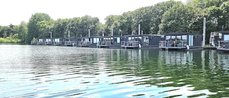 A row of floating houseboats with outdoor seating areas and windows, docked along a river.