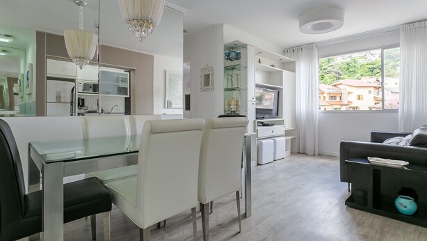 A modern living room with a glass dining table, white chairs, a black sofa, and a large window with curtains. - WI-FI 100MB | 200m from Beiramar Shopping #CA44D (Florianópolis)