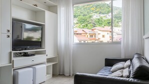 A modern living room with a flat-screen TV, a sofa, and a view of a hillside town through a large window. - WI-FI 100MB | 200m from Beiramar Shopping #CA44D (Florianópolis)
