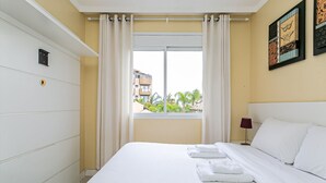 Une chambre d’hôtel avec un grand lit, des rideaux blancs, une fenêtre donnant sur des bâtiments et de la verdure, et un luminaire fixé au mur.