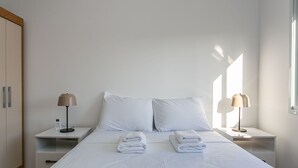 En pænt redt seng med hvide sengetøj og to sammenfoldede håndklæder. To sengeborde, hver med en lampe og en telefon. Væggene er hvide, og der er et træskab.