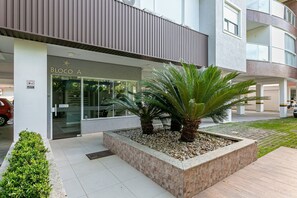 A modern building with a glass entrance, a landscaped area with palm trees, and a sign reading "BLOCO A". - 500m from beach | Waterfront gastronomy #CHA05D (Florianópolis)
