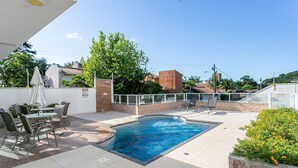 En swimmingpool med fliseterrasse, omgivet af stole og et bord, med udsigt til bygninger og træer i baggrunden.