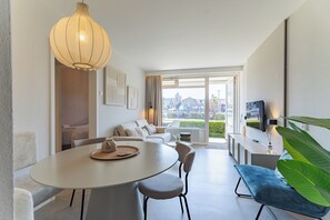 A modern living room with a dining area, a large window with a city view, and a television mounted on the wall.