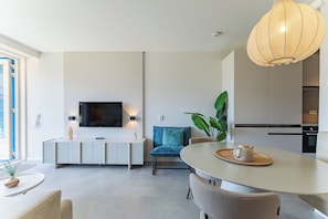 A modern living room with a flat-screen TV, a dining area with a round table, and a kitchen area with a stove.