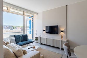 Un salon moderne avec un canapé, un fauteuil bleu, une table basse, un téléviseur fixé au mur et une vue sur l’extérieur.