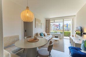 A modern living room with a dining table, sofa, and chairs, a hanging light fixture, and a large window with a view of the outside.