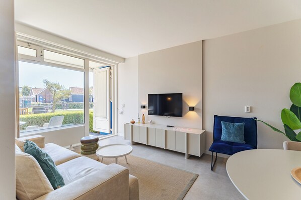 A modern living room with a sofa, a round table, a TV mounted on the wall, and a large window.