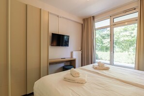 A hotel room with a bed, a TV mounted on the wall, a window with curtains, and a small table with a remote control.