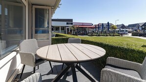Eine Terrasse mit Korbmöbeln, einem runden Tisch und Blick auf ein Gewerbegebiet mit Geschäften und einem Parkplatz.