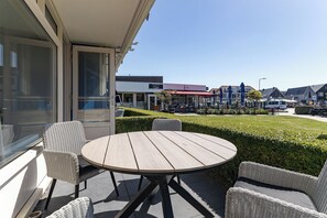 A patio with wicker furniture, a round table, and a view of a commercial area with shops and a parking lot.