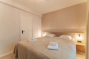A hotel room with a bed, a door, a nightstand with a lamp, and a wall-mounted light fixture.