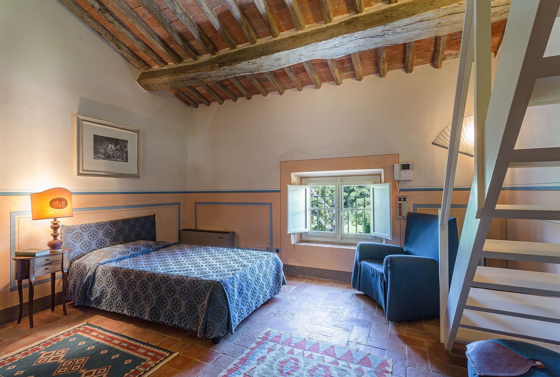 A bedroom with a bed, a chair, a window, a lamp, and a staircase.