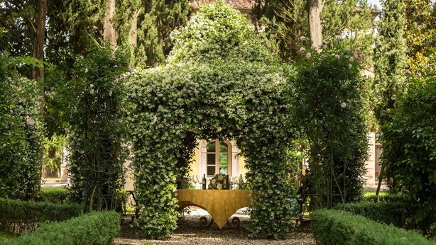 A garden with a central archway surrounded by lush greenery and a table set for a meal.