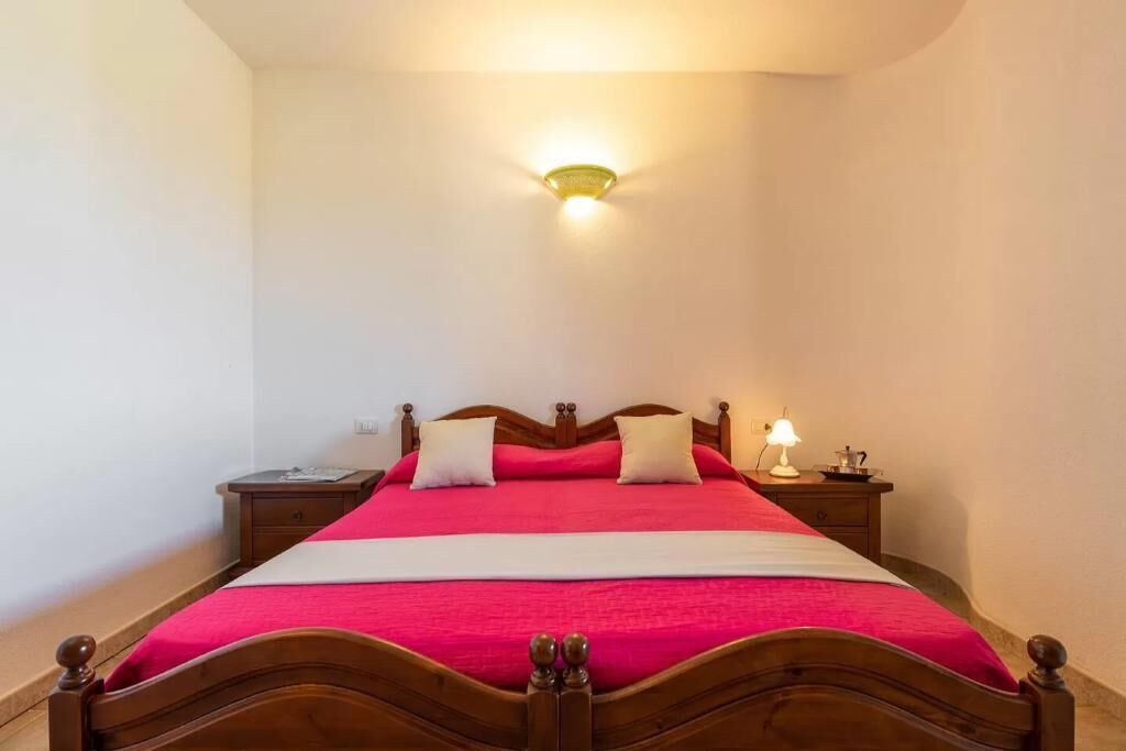 Ein ordentlich bezogenes Bett mit Holz-Kopfteil, ein Nachttisch mit Lampe und eine Wandleuchte.