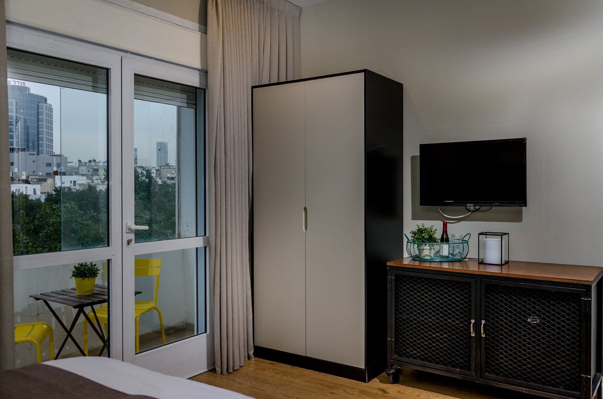 Une chambre d’hôtel avec une grande fenêtre, une armoire, un téléviseur, une petite table avec une plante et une chaise.