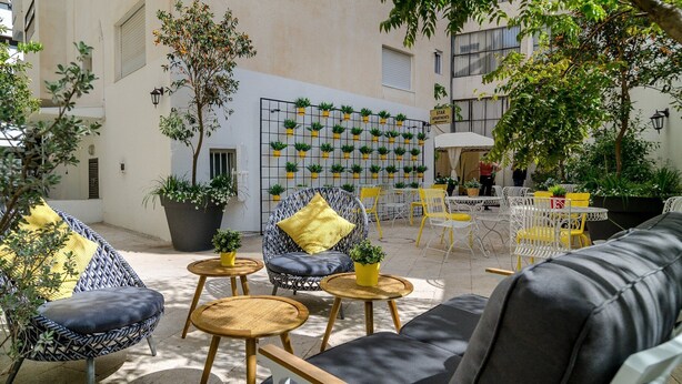 An outdoor seating area with wicker chairs and a sofa, surrounded by potted plants and a wall with hanging planters. - Dizengoff square boutique (Tel Aviv)