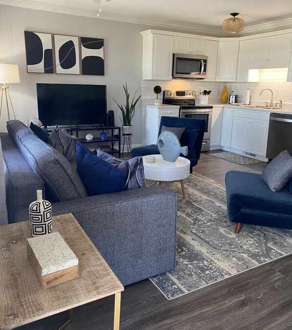 A modern living room with a grey sectional sofa, a blue armchair, and a coffee table. A flat-screen TV and a small side table with a decorative vase are also visible.
