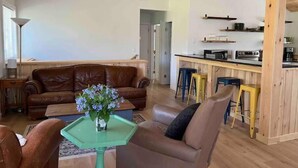 A spacious living area with a brown leather sofa, a green table with a vase of flowers, and a kitchen area with stools and appliances. - River Retreat with Kayaks, Firepits, Stars (Strasburg)